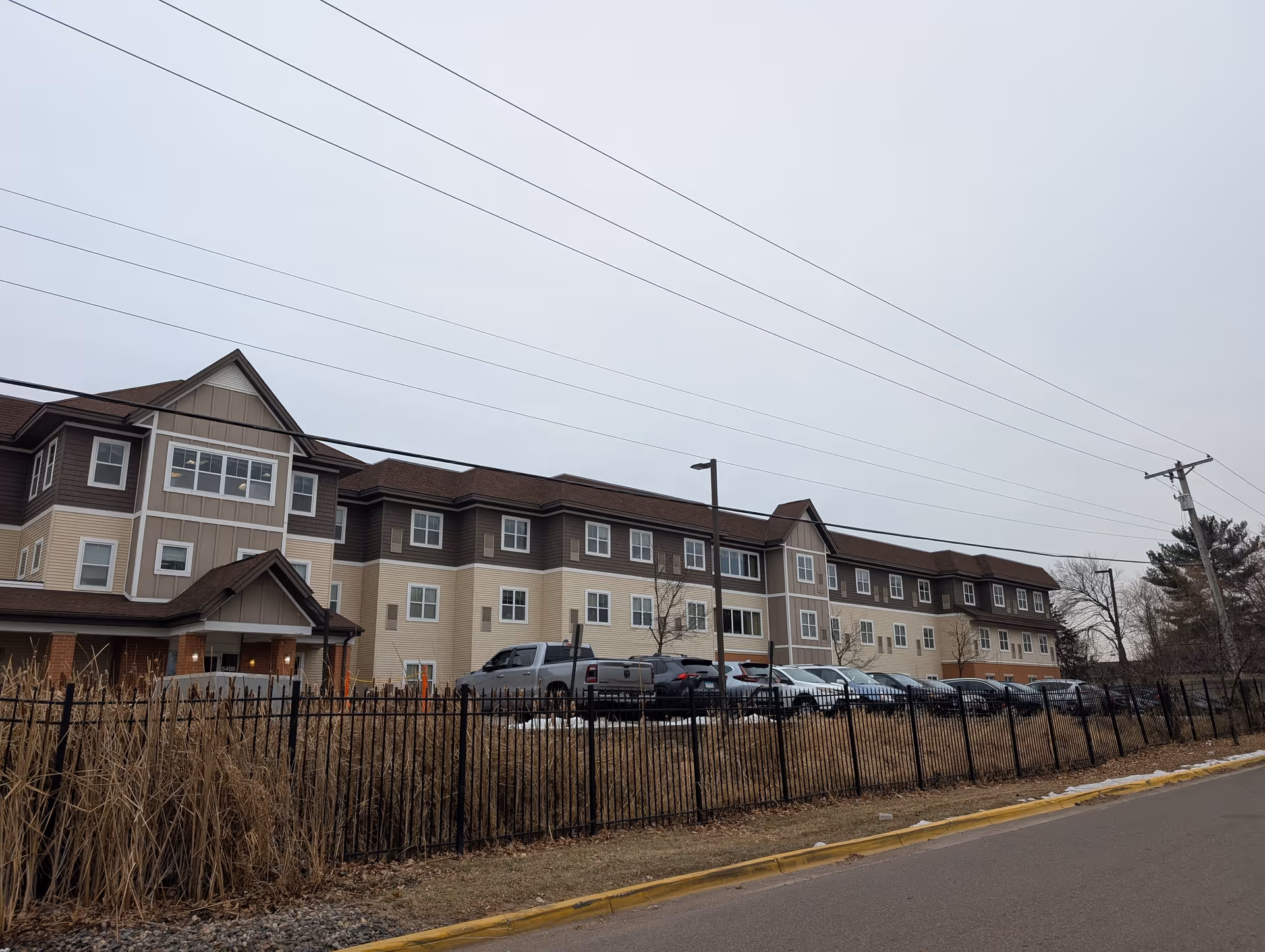 Exterior front view of a three-story senior living building with parked cars, a black metal fence, and overhead power lines.