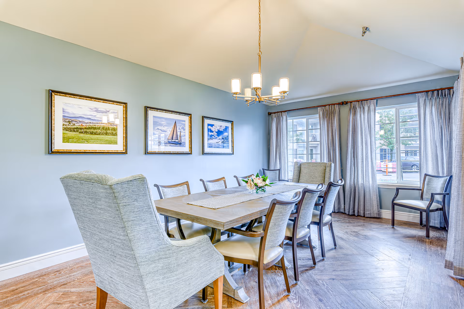 Bright dining room with a wooden table surrounded by upholstered chairs, a chandelier overhead, framed artwork on the wall and large windows with curtains.