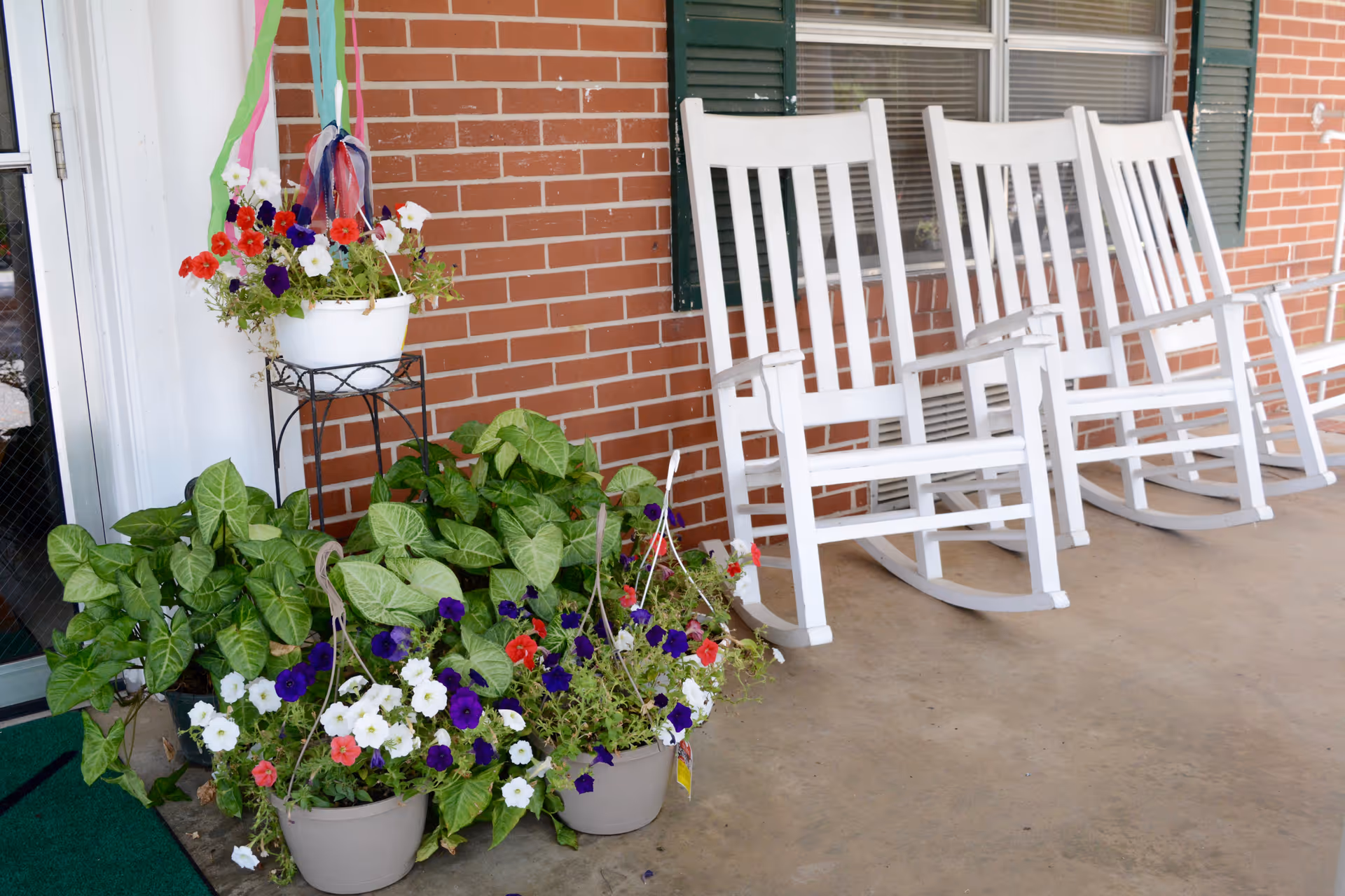 A porch area with three white wooden rocking chairs lined up against a brick wall with green shutters. Several pots of colorful flowers and green plants are placed near the chairs, including a hanging white pot with red, purple, and white flowers. The porch floor is concrete.