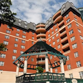 View looking up at a multi-story red brick building with numerous windows and balconies, featuring a green gazebo structure in the foreground under a partly cloudy sky.