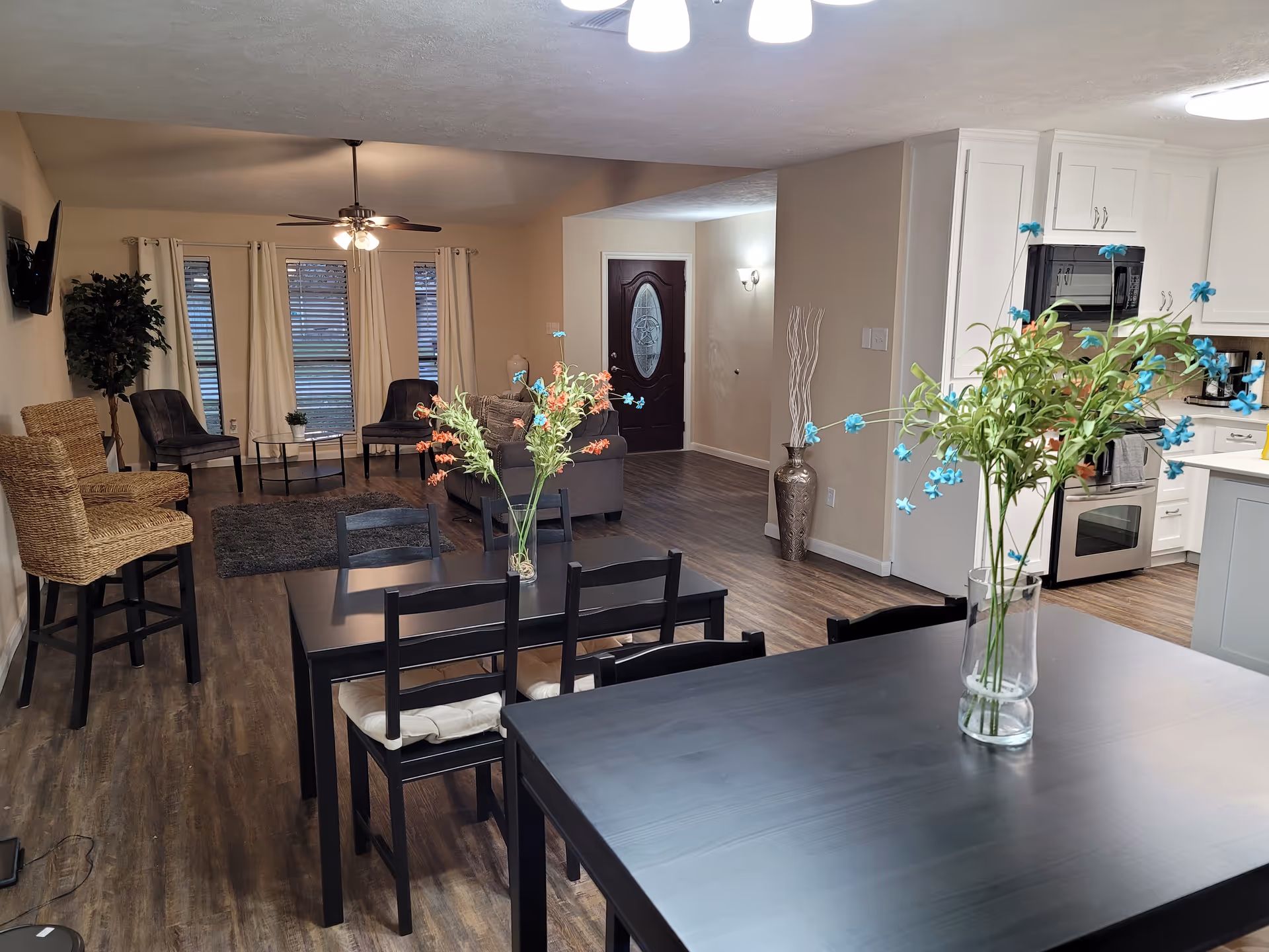 Interior view of a senior living facility showing a combined dining and living area. In the foreground, there is a black dining table with chairs and a vase with blue flowers. Behind it, another dining table with chairs and a vase with orange flowers is visible. The living area has a ceiling fan, a sofa, two chairs, a small round table, and a potted plant near the windows with white curtains. The kitchen area with white cabinets, a microwave, and an oven is visible on the right side. The entrance door is seen in the background.