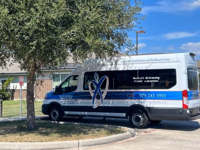 A white and blue shuttle van parked near a tree outside a building. The van has the logo and name 'Arbor Hills Rehabilitation and Healthcare Center' on its side along with a phone number and website. The background shows part of a brick building and a clear blue sky.