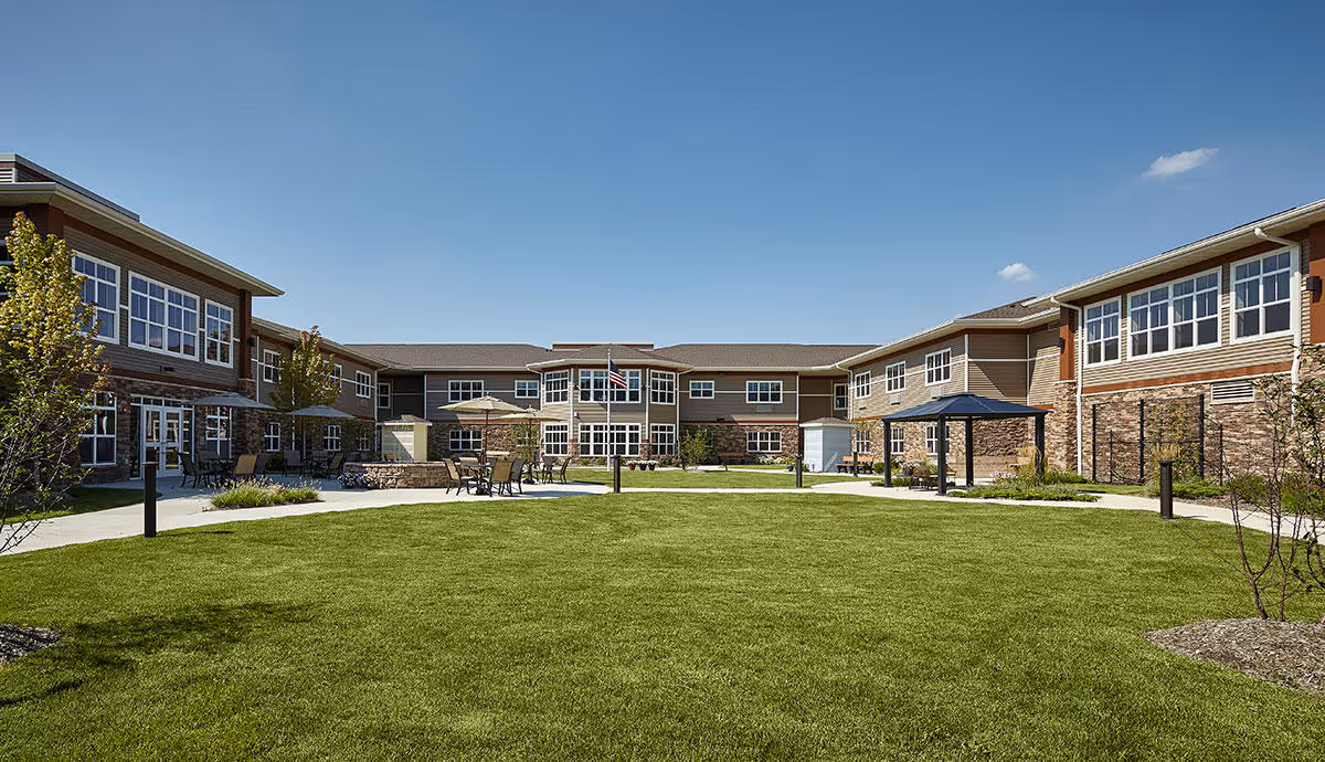 Outdoor courtyard area of Terra Vista of Oakbrook Terrace featuring a large green lawn surrounded by a two-story building with many windows. There are several patio tables with umbrellas and chairs on paved walkways around the lawn, under a clear blue sky.