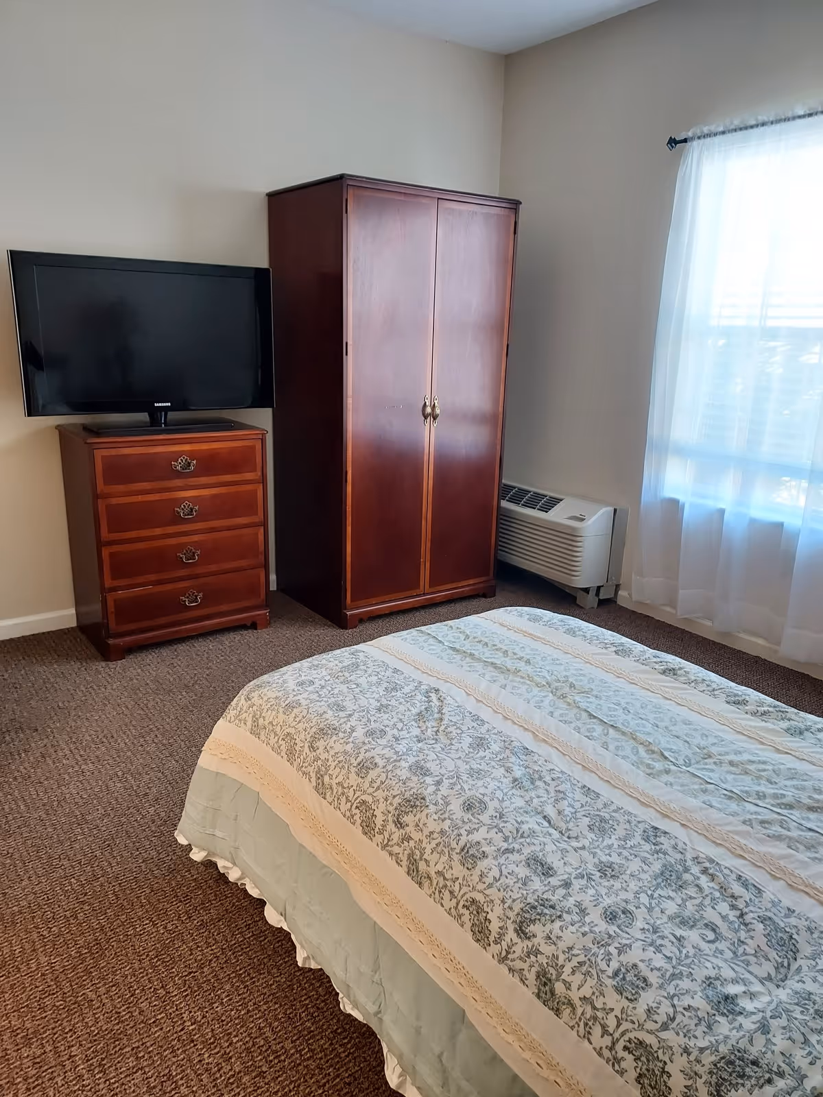 A bedroom with a bed covered in a patterned quilt, a wooden dresser with a flat-screen TV on top, a wooden wardrobe, and a window with sheer white curtains allowing natural light to enter. There is a wall-mounted air conditioning unit below the window.