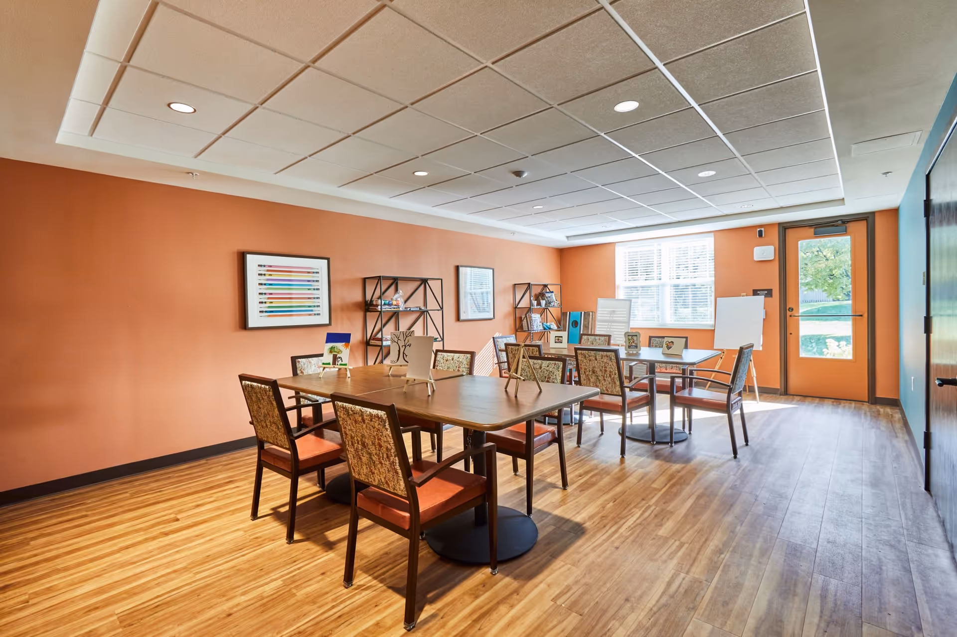 A well-lit room with orange walls and wooden flooring featuring two tables surrounded by chairs with patterned upholstery and orange cushions. The tables display small easels holding artwork. There are two black metal shelving units against the wall with various items on them. A large window and a glass door allow natural light to enter the room.
