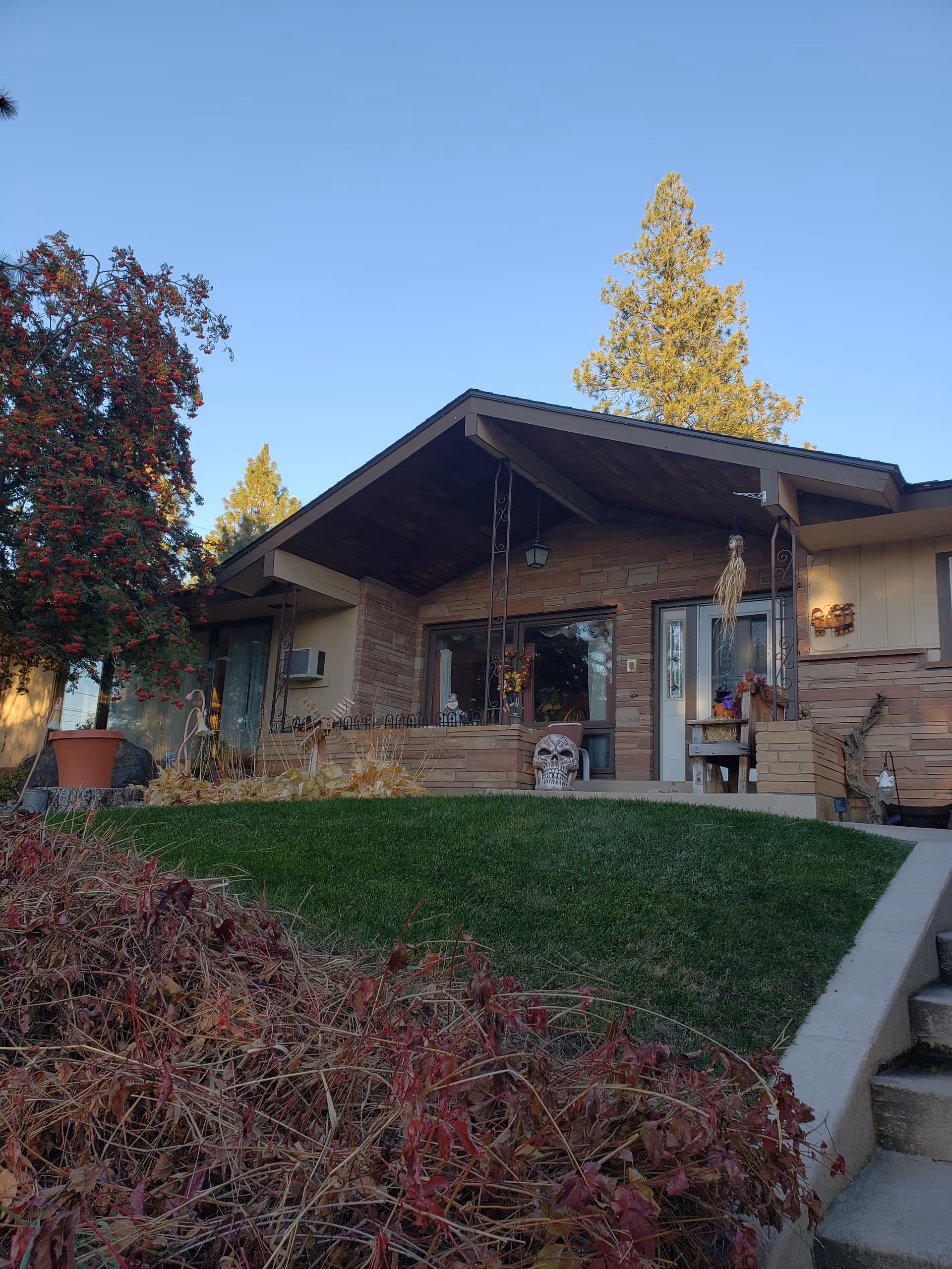 Front exterior view of a single-story house with a sloped roof, a small porch decorated with autumn-themed items including a large skull decoration, and a well-maintained green lawn with some dry plants in the foreground. There are tall trees and a clear blue sky in the background.