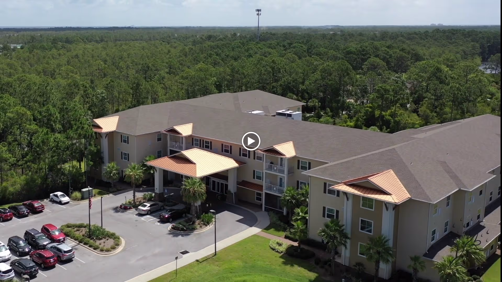 Aerial view of a large, three-story senior living facility building with a covered entrance, surrounded by a parking lot with several cars and lush green trees in the background under a partly cloudy sky.