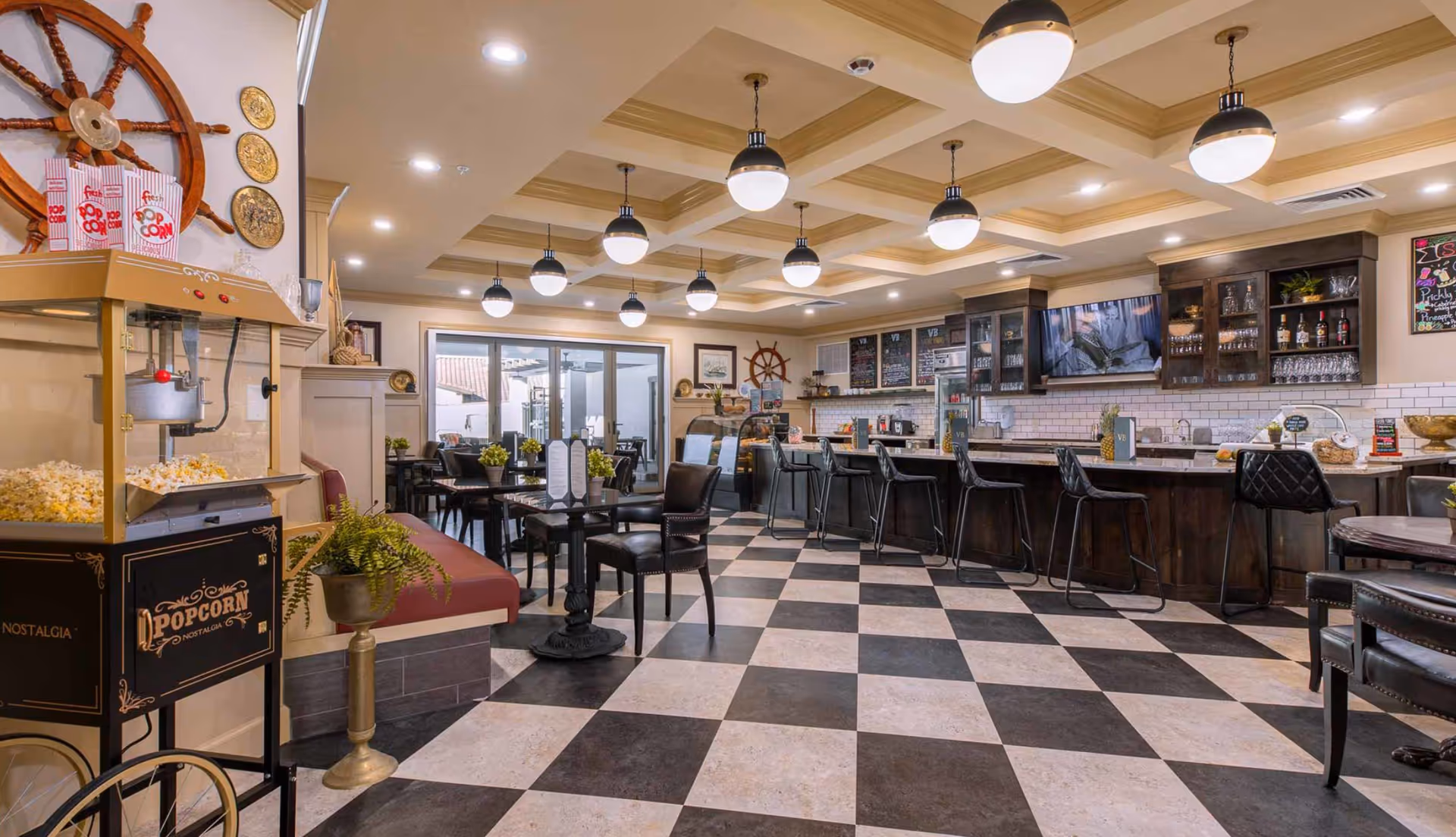 Communal dining and bar lounge with a black-and-white checkered floor, round tables and bar seating, pendant lights, and a popcorn machine.