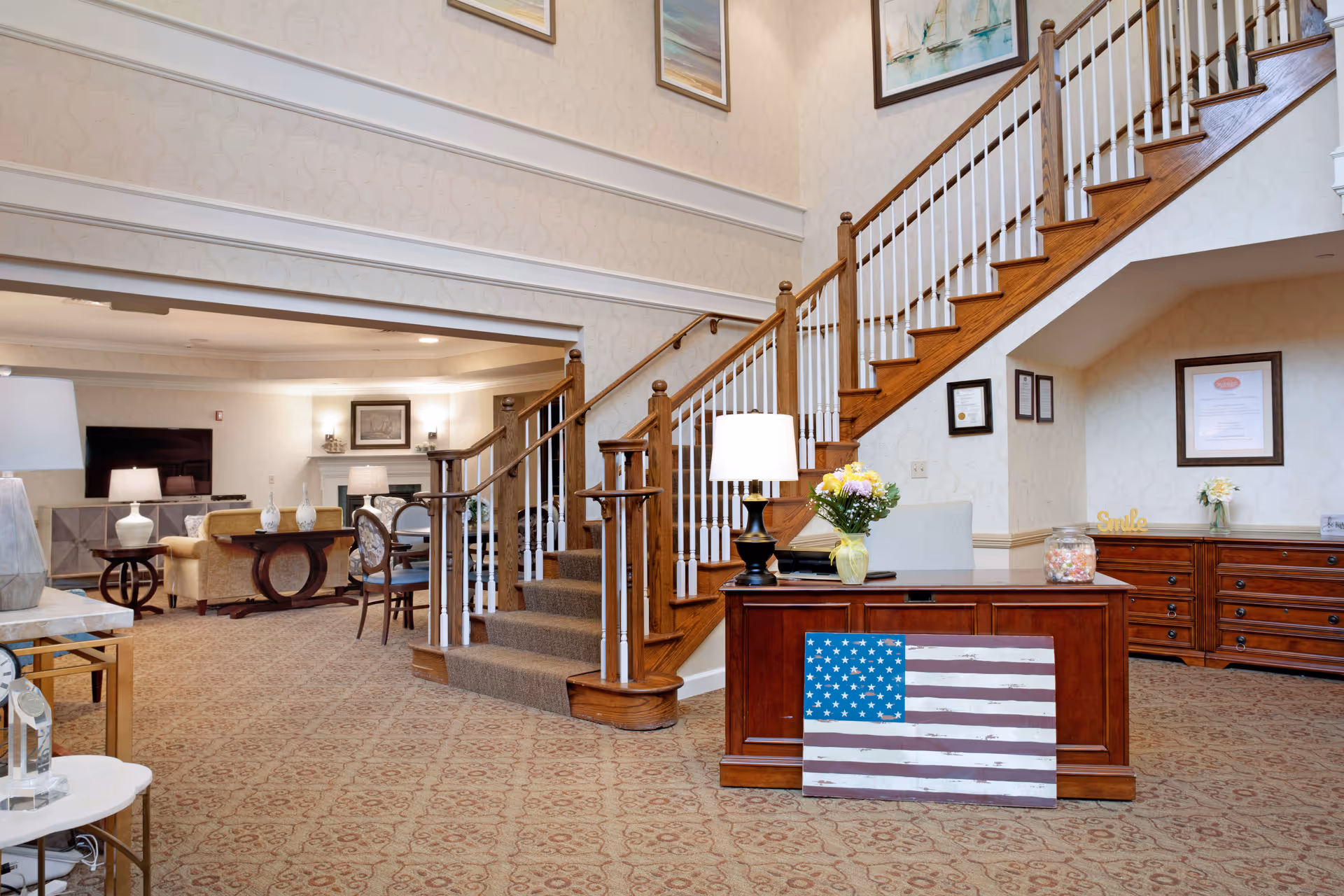 A bright senior living lobby with a wooden staircase, reception desk displaying an American flag artwork, and a seating area with lamps and decor.