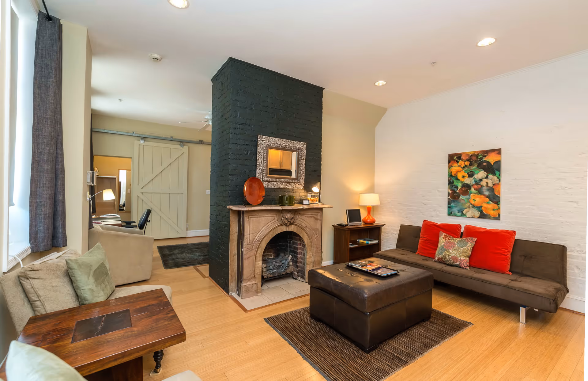 A cozy living room with a dark brown sofa adorned with red and patterned cushions, a large dark brown ottoman on a brown rug, a stone fireplace with a black painted chimney and a decorative mirror above it, a wooden side table with a lamp and books, light wood flooring, and a colorful painting on the white brick wall.