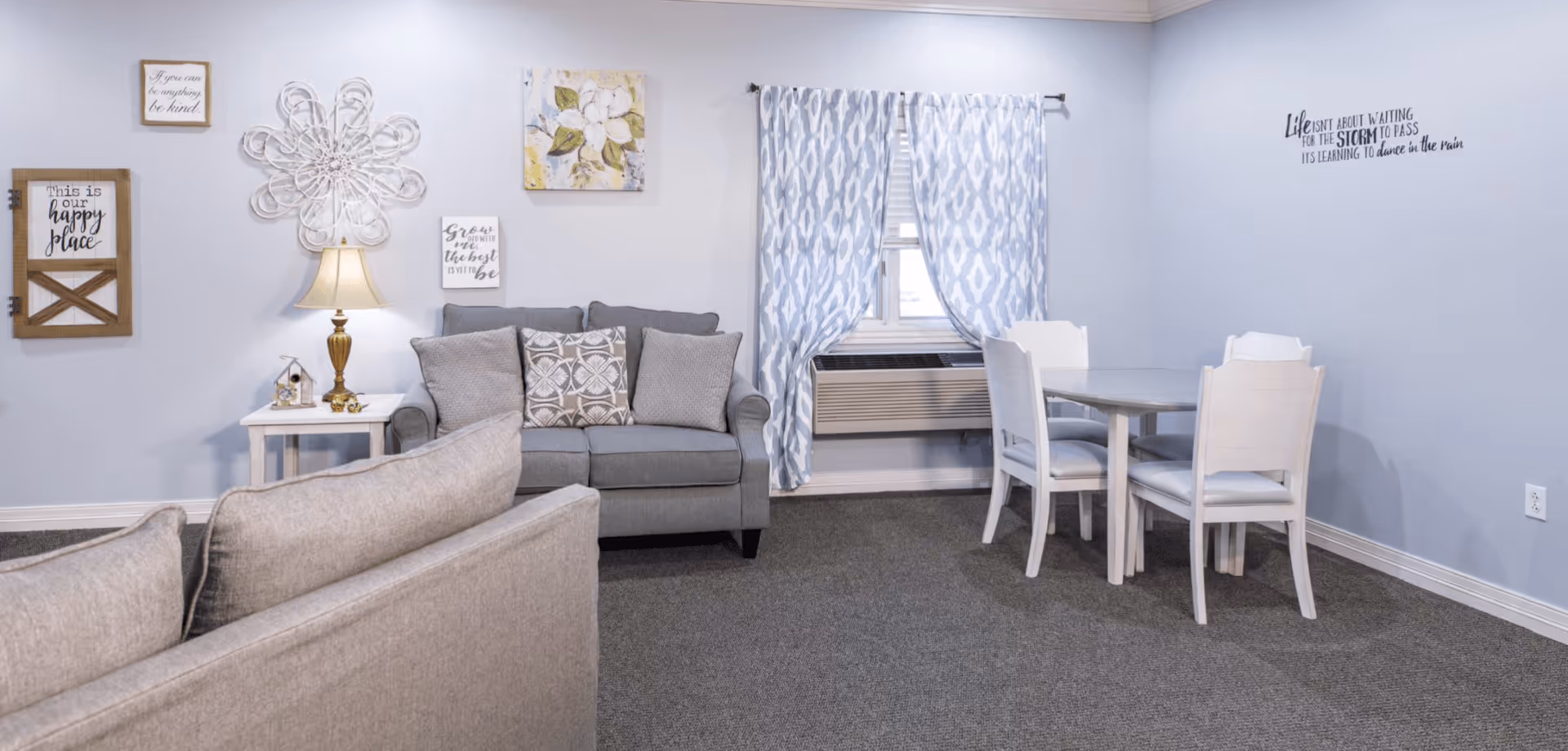 A cozy living area with light blue walls featuring decorative wall art and quotes. There are two gray sofas with patterned cushions, a small white side table with a lamp, and a white dining table with four chairs near a window with blue and white patterned curtains. An air conditioning unit is installed below the window.