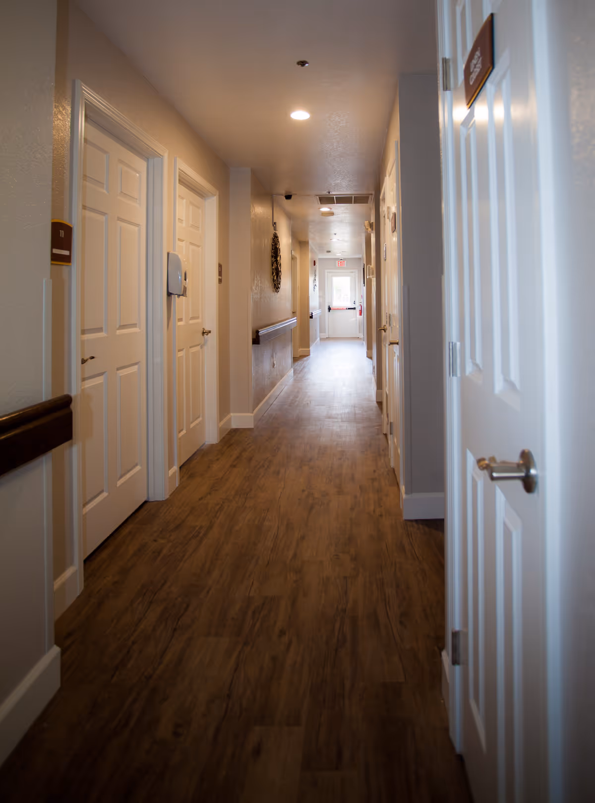 A long, well-lit hallway in a senior living facility with wooden flooring and beige walls. Several white doors line both sides of the hallway, some with room number signs. Handrails are mounted on the walls, and there is a decorative wall piece on the left side. At the end of the hallway, there is a door with an exit sign above it.