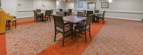 A spacious room with several square tables and chairs arranged for seating. The floor is covered with a patterned carpet in shades of red and beige. The walls are light-colored with framed pictures and a bulletin board. A piano is visible on the left side of the room.