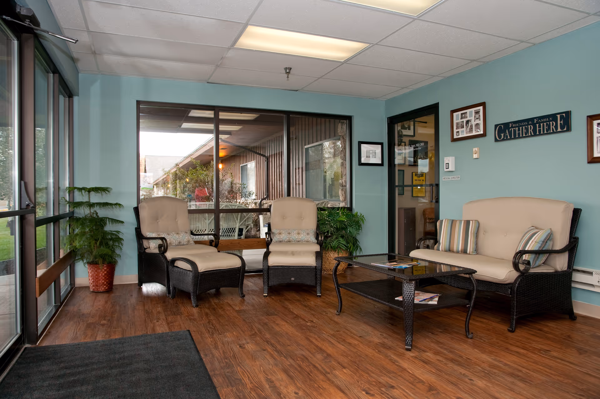 A cozy seating area in a facility with light blue walls and wood flooring. The space includes two cushioned armchairs with matching ottomans, a cushioned loveseat with striped pillows, and a glass-top coffee table. There are framed pictures and a sign on the wall that reads 'Friends & Family Gather Here.' Large windows and a glass door allow natural light to enter, and there are potted plants adding greenery to the room.
