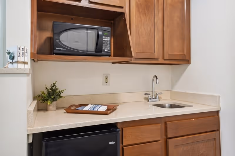 A small kitchenette area featuring wooden cabinets, a black microwave on an upper shelf, a small sink with a chrome faucet, a black mini refrigerator below the counter, a small potted plant, and a tray with a folded cloth on the countertop.