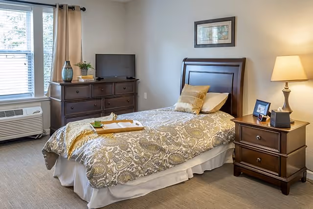 A cozy bedroom with a single bed covered in a patterned comforter and pillows. Next to the bed is a wooden nightstand with a lamp, a framed photo, and a small box. Across from the bed is a wooden dresser with a small TV, a plant, and decorative items. A window with curtains lets in natural light, and an air conditioning unit is below the window.