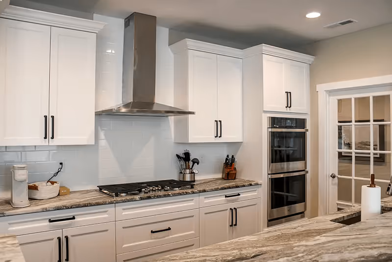 Modern kitchen with white cabinets, a stainless steel range hood, a built-in double oven, a gas cooktop, and a marble countertop. There is a glass-paneled door on the right side and kitchen utensils on the countertop.