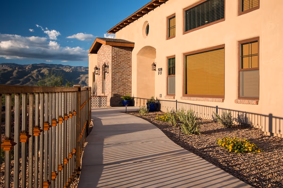 Exterior view of a building at The Villas at Houghton with a curved concrete walkway, metal railing, desert landscaping with small plants and flowers, and mountains in the background under a partly cloudy sky.