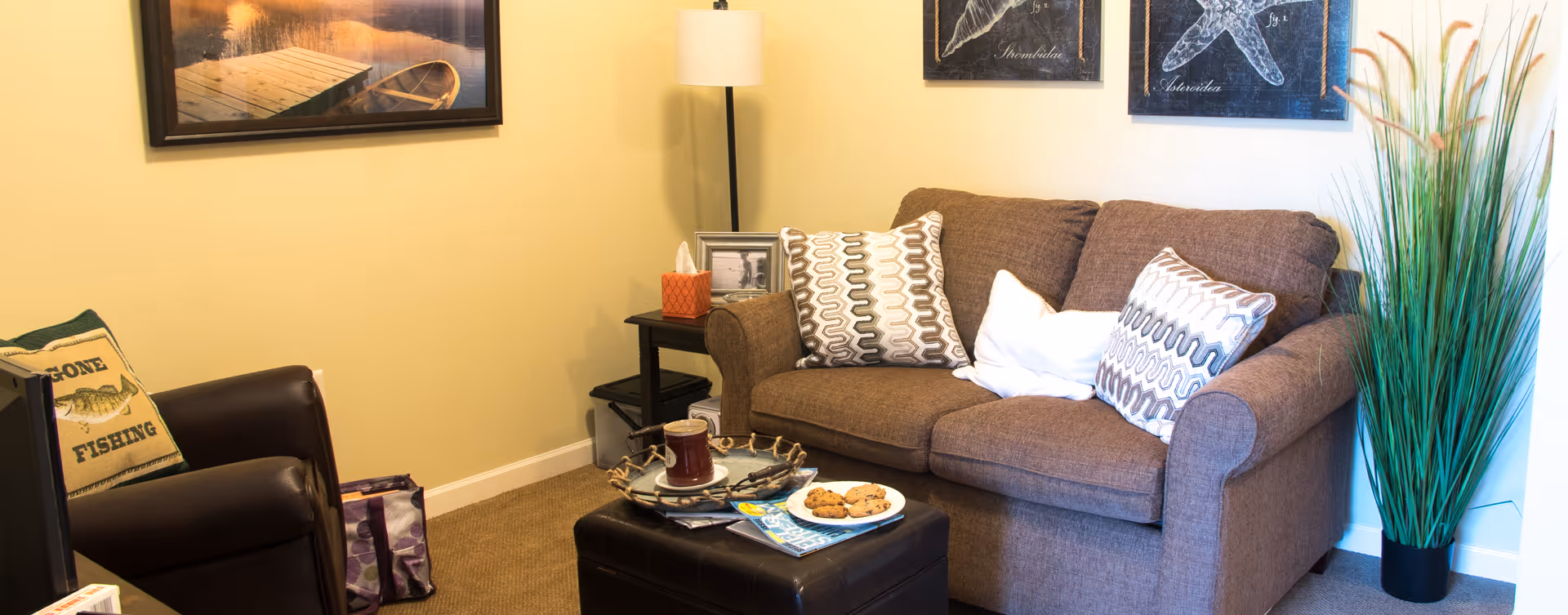 Cozy living room featuring a brown loveseat with patterned pillows, an armchair and ottoman with snacks, a side table and floor lamp, and a tall potted plant.