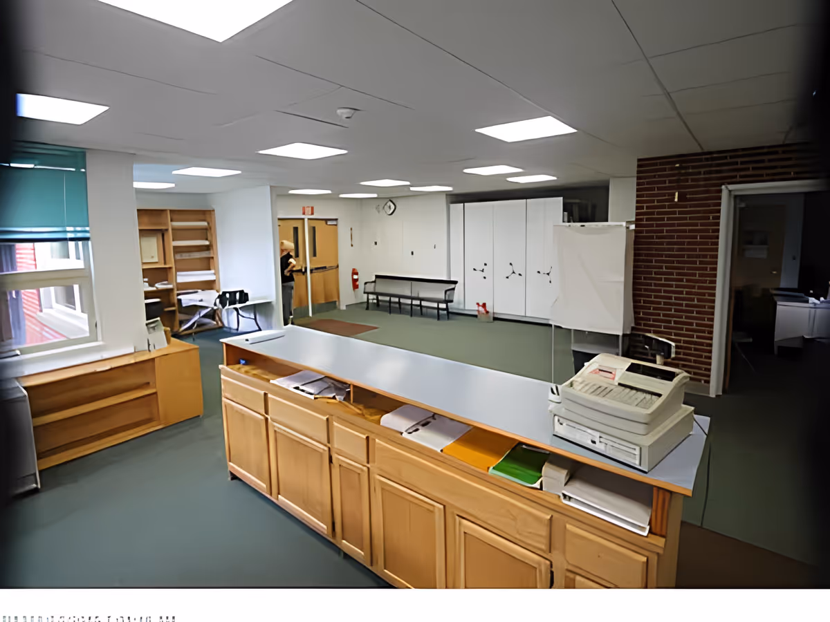 Interior view of a facility room with a long wooden counter in the foreground holding papers and a fax machine. The room has green flooring, white walls, and a ceiling with fluorescent lights. There are shelves, a bench, a fire extinguisher, and a clock on the wall. A person is partially visible near a door in the background.