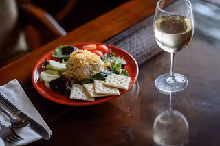 A red plate with a serving of salad topped with shredded cheese, cherry tomatoes, and several saltine crackers on a wooden table next to a glass of white wine. A napkin with a fork and spoon is placed to the left of the plate.
