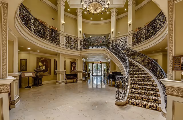 Grand two-story lobby with an ornate curved staircase, decorative iron railings, chandelier, and marble floors.