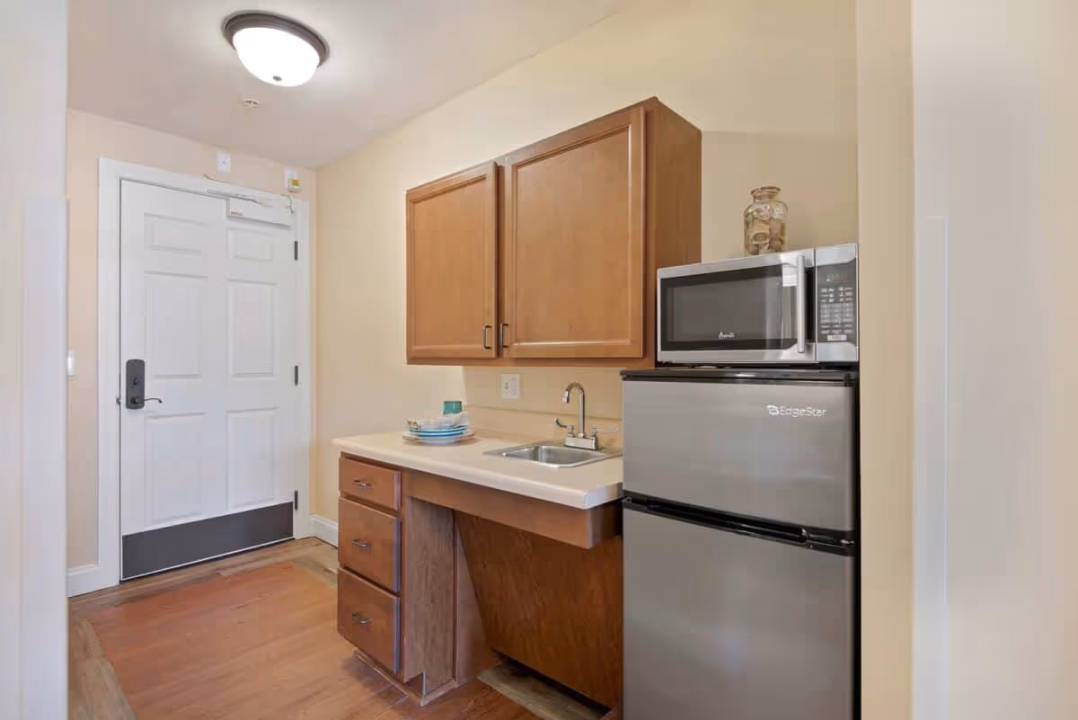 Small kitchenette area with a white door on the left, wooden cabinets above and below a countertop with a small sink. A stack of plates and a cup are on the counter. To the right, there is a compact stainless steel refrigerator with a microwave on top and a glass jar filled with corks.