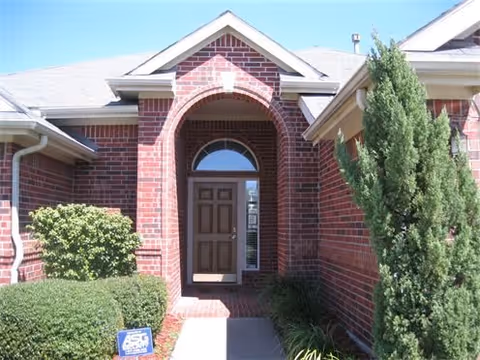 Front entrance of a brick residential building with an arched doorway, a brown door, and surrounding greenery including bushes and a tall tree.