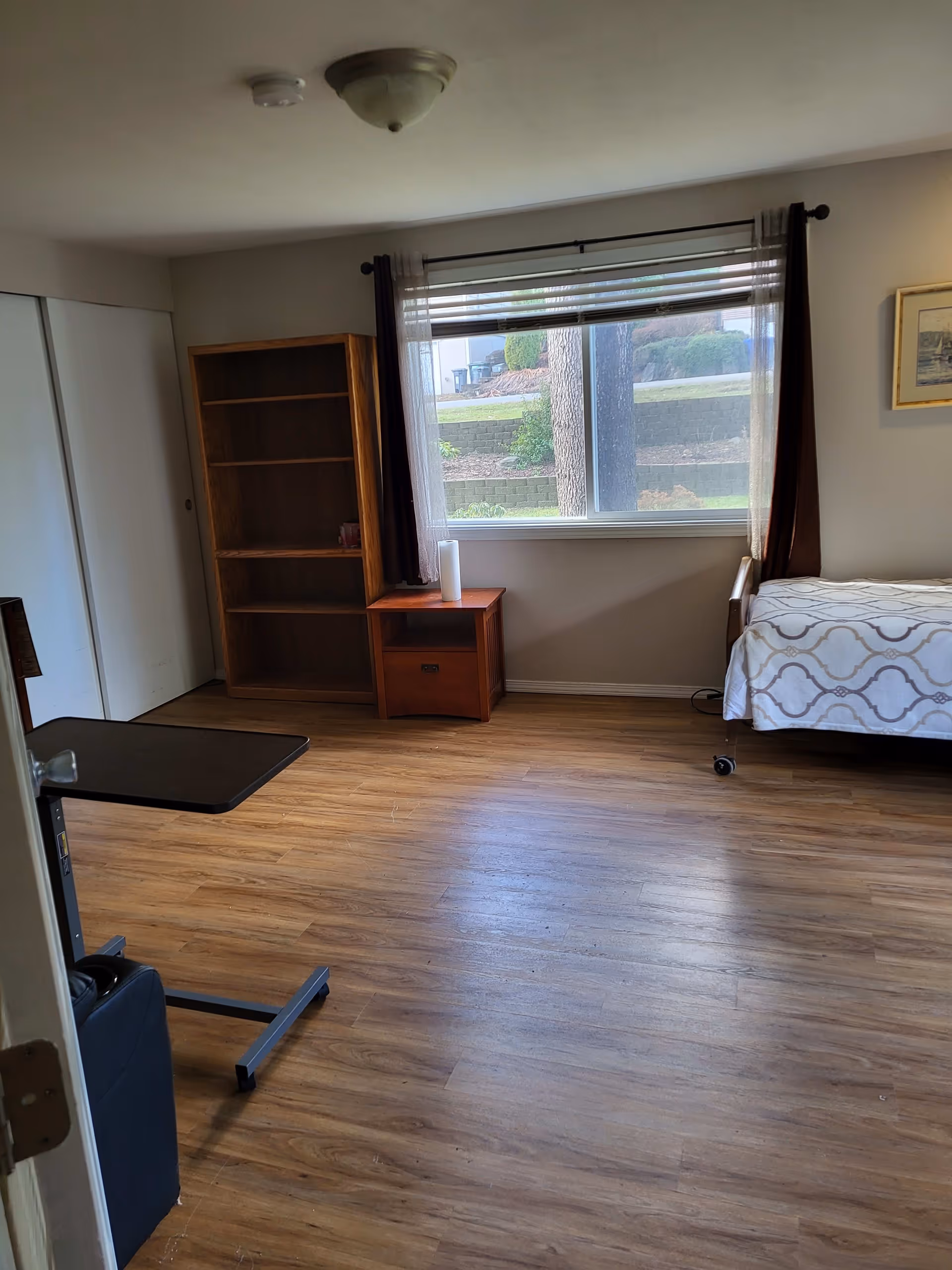 A simple bedroom with wooden flooring, a single bed with patterned bedding on the right, a wooden bookshelf and a small wooden nightstand with a white cylindrical object on it near a window with brown curtains and white blinds. The room has white walls and a ceiling light fixture.