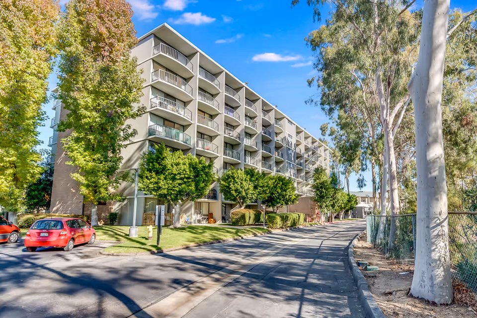 Exterior view of a multi-story senior living facility building with balconies, surrounded by trees and a driveway with parked cars under a clear blue sky.