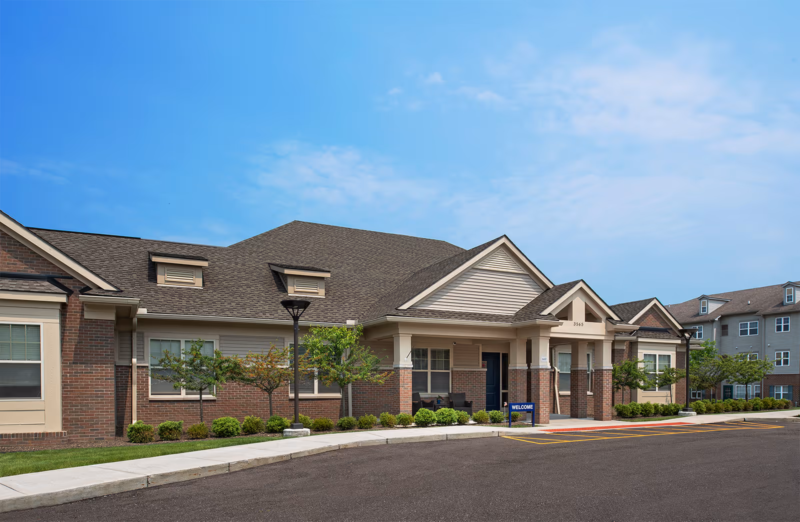 Exterior view of a single-story brick and siding building with a covered entrance and a 'Welcome' sign near the door, surrounded by small trees and shrubs under a blue sky.