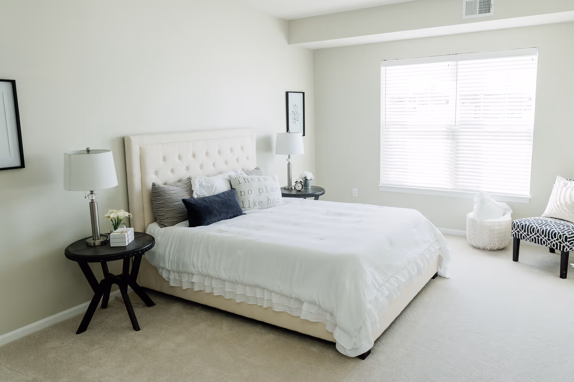 A bright and clean bedroom with a large bed featuring a tufted beige headboard, white bedding, and multiple pillows including one with the text 'There's no place like home'. There are two round black bedside tables each with a lamp, a small plant, and a clock. A window with white blinds lets in natural light, and a patterned chair and a basket with a blanket are placed near the window.