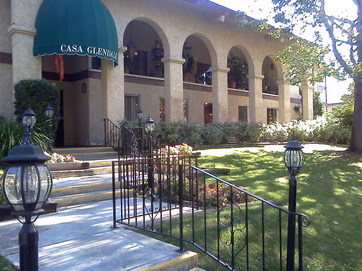 Exterior view of a building with beige stucco walls and arched windows. There is a green awning above the entrance with the text 'CASA GLENDALE'. A pathway with black wrought iron railings and lamp posts leads to the entrance. The surrounding area has green grass, bushes, and trees.