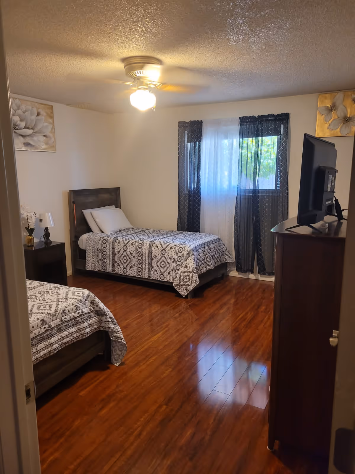 A bedroom with two single beds covered in patterned bedspreads. There is a wooden nightstand with a lamp and a small plant between the beds. A ceiling fan with a light is on the ceiling. The room has wooden flooring, a window with sheer and dark curtains, and a wooden dresser with a TV on top. The walls are decorated with floral artwork.