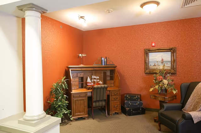 Cozy sitting area with a wooden roll-top desk, striped chair, armchair, decorative column, and framed artwork against red patterned wallpaper.
