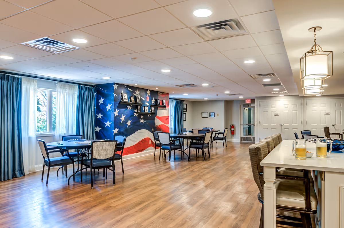 A spacious common area with wooden flooring and multiple tables and chairs arranged for seating. One wall features a large mural of the American flag with shelves displaying framed photos. Large windows with blue curtains allow natural light to enter the room. There is a countertop with bar stools and two glasses of beer on it. The ceiling has recessed lighting and hanging light fixtures.