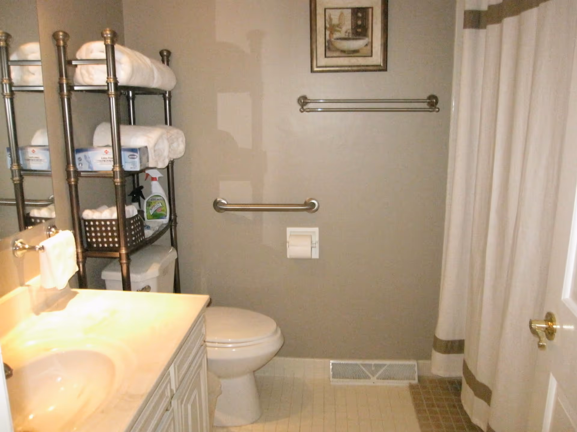 A bathroom with a white sink and countertop on the left, a toilet in the center, and a metal shelving unit above the toilet holding folded white towels, cleaning supplies, and a basket. There is a towel rack and a framed picture on the wall above the toilet, and a shower curtain on the right side.