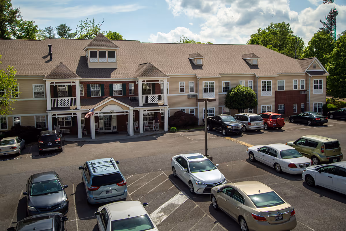 Front exterior of a two-story senior living building with balconies, a covered entrance, and a parking lot with cars.