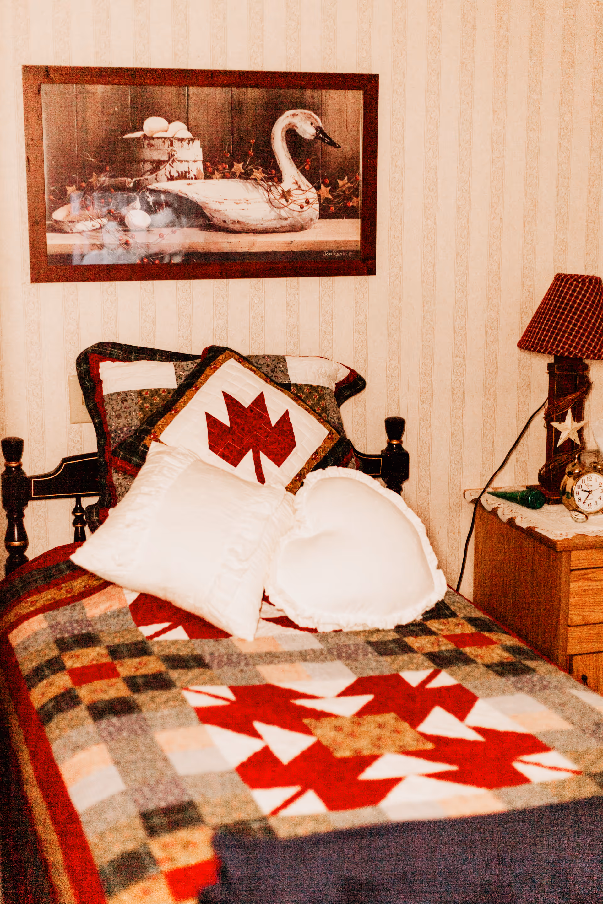 A cozy single bed with a colorful patchwork quilt featuring red, white, and green patterns. The bed has two decorative pillows, one square and one heart-shaped, both white. Above the bed hangs a framed picture of a white swan with eggs and autumn leaves. To the right of the bed is a wooden nightstand with a red checkered lampshade, an alarm clock, and a small green object. The wall behind the bed has a light patterned wallpaper.