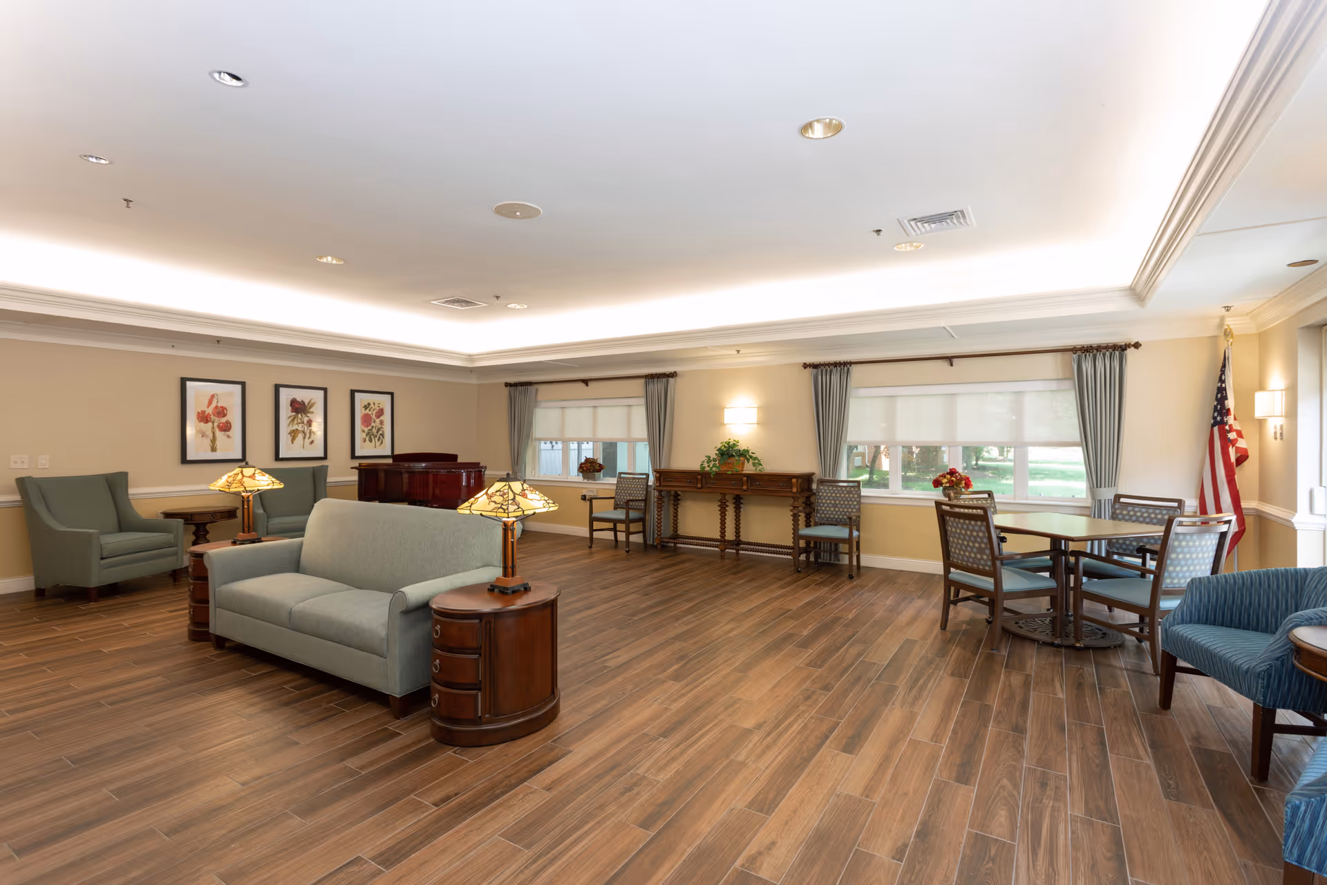 A spacious, well-lit common area with wooden flooring, light-colored walls, and a white ceiling with recessed lighting. The room features a light green sofa with two matching table lamps on wooden side tables, two green armchairs, a wooden console table with plants and flowers, a round table with four chairs, and large windows with gray curtains letting in natural light. An American flag stands in the corner near the windows.