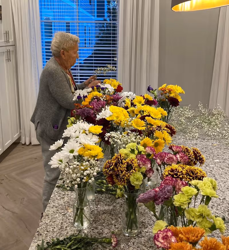 An elderly woman arranging multiple colorful flower bouquets on a large granite countertop in a well-lit kitchen with white cabinets and a window with blinds in the background.