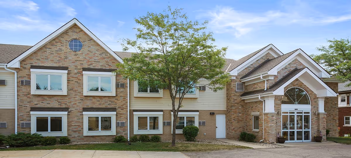 Exterior view of a two-story senior living facility building with brick and siding facade, multiple windows, a central tree, and a covered entrance with glass doors under a gabled roof.