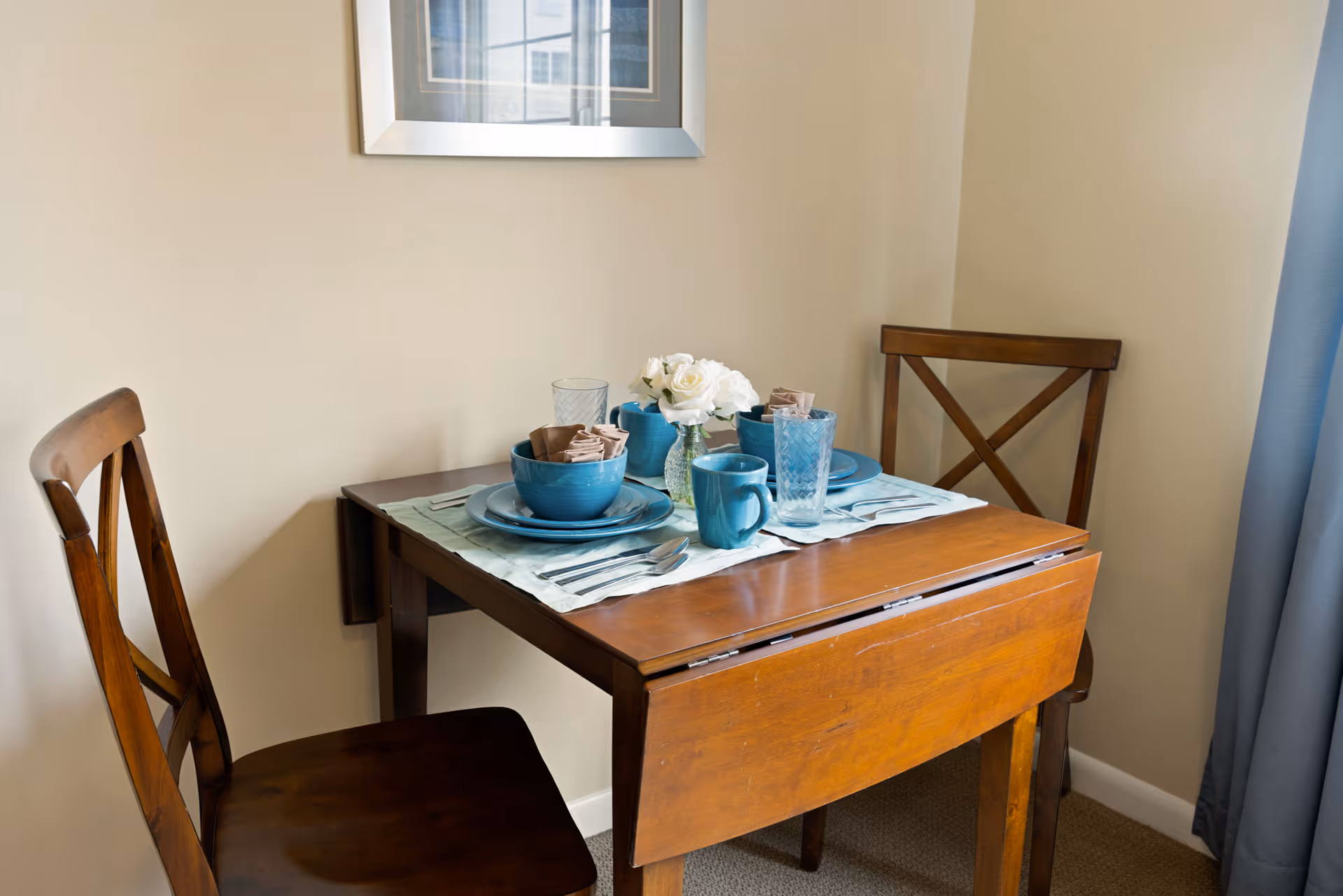 A small wooden dining table set for two with blue dishes, mugs, and glasses, along with a small vase of white flowers. Two wooden chairs are positioned on either side of the table. A framed picture hangs on the beige wall above the table, and a blue curtain is partially visible on the right side.