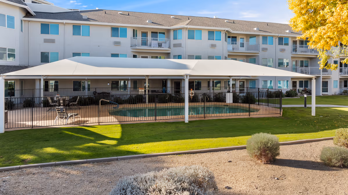 Outdoor view of a senior living community building with a covered swimming pool area enclosed by a black metal fence. The building is three stories tall with multiple windows and balconies. There is a well-maintained lawn and some desert landscaping with bushes and a tree with yellow leaves in the foreground.