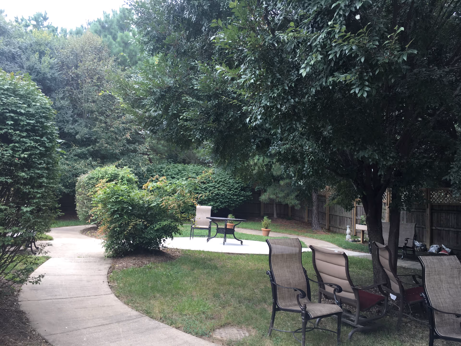 Outdoor garden area with a curved concrete pathway, surrounded by green bushes and trees. Several patio chairs and a small table are arranged on the grass near a large tree. A wooden fence encloses the area.