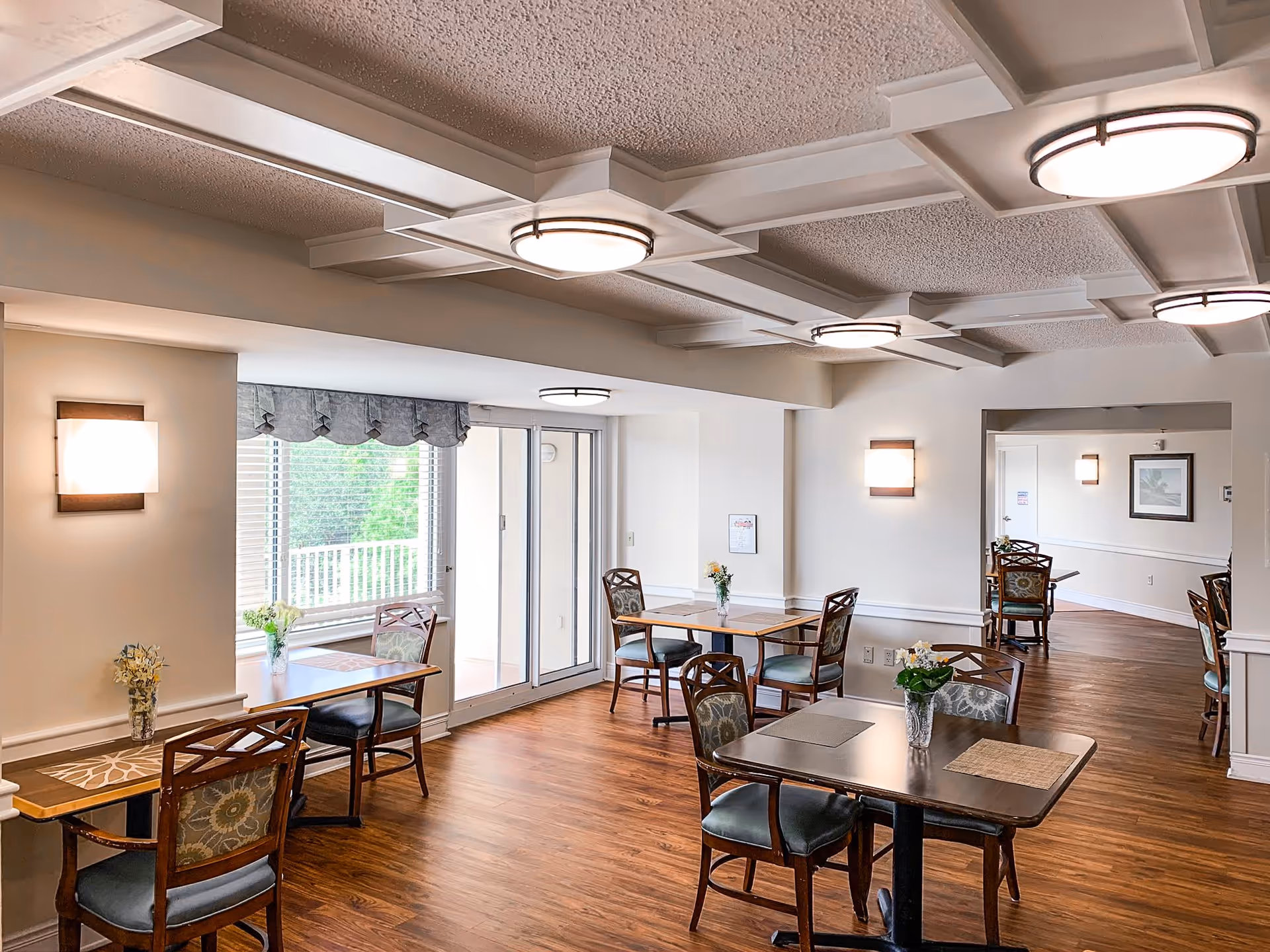 A bright and clean dining area with wooden floors, several tables with chairs, and small flower vases on each table. The room has large windows with blinds and a sliding glass door leading outside. The ceiling features decorative beams and round light fixtures, and the walls are painted white with wall-mounted lights.
