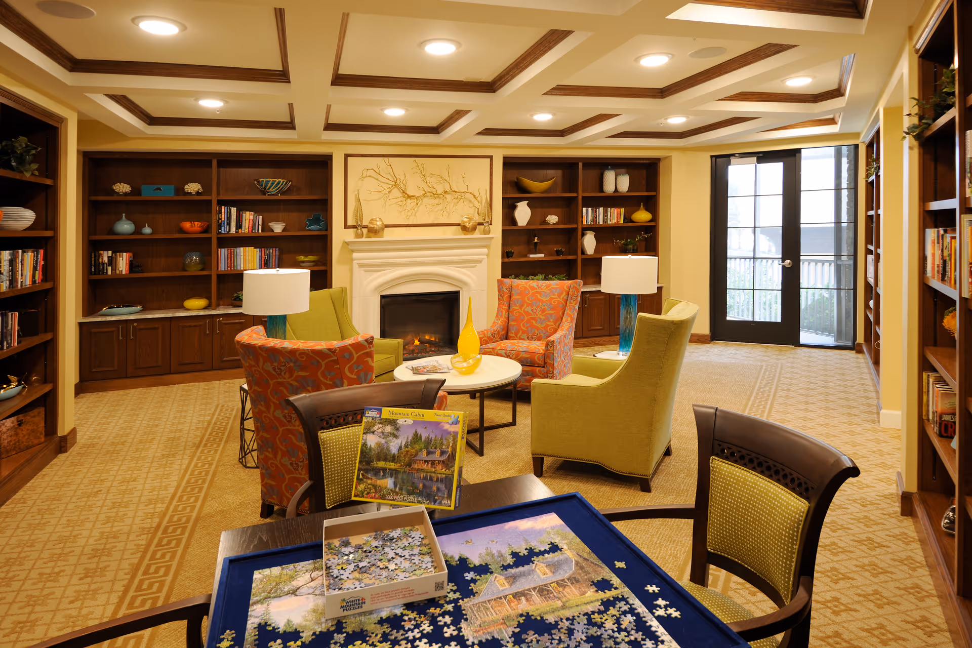 A cozy senior living common area with a puzzle table in the foreground, partially completed puzzle and box on it. The room features comfortable armchairs in green and orange patterns arranged around a small round table with a yellow vase. Behind the seating area is a fireplace with decorative art above it, flanked by built-in wooden bookshelves filled with books and decorative items. Large glass doors lead outside, and the room has warm lighting and a patterned carpet.