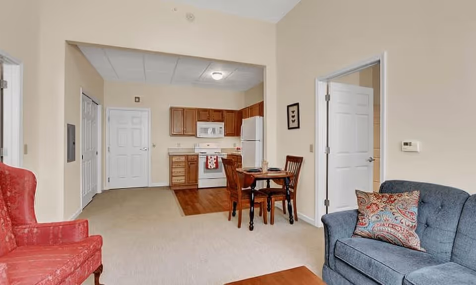 Interior view of a senior living facility apartment showing a small kitchen with wooden cabinets, white appliances, and a dining table with two chairs. The living area has a red armchair and a blue sofa with a decorative pillow. The walls are painted beige and there are two white doors visible.