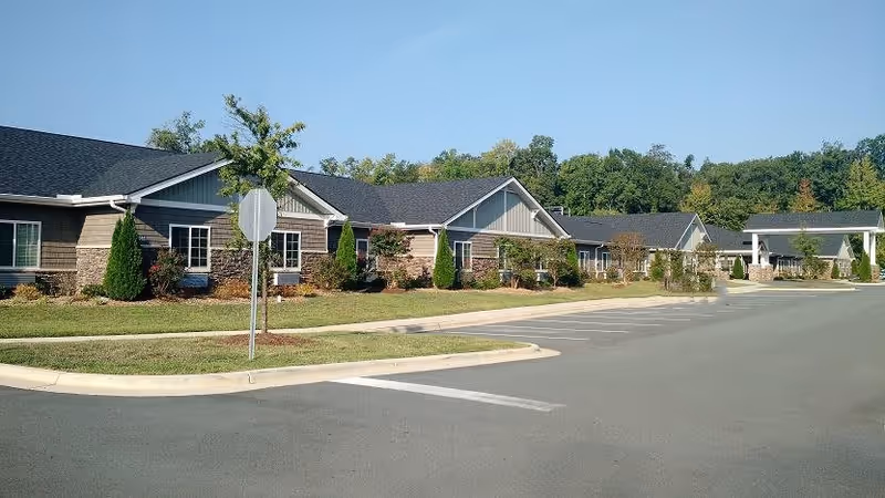 Exterior view of a single-story building with a combination of stone and siding facade, surrounded by landscaped bushes and trees, with a parking lot in the foreground and a clear blue sky above.