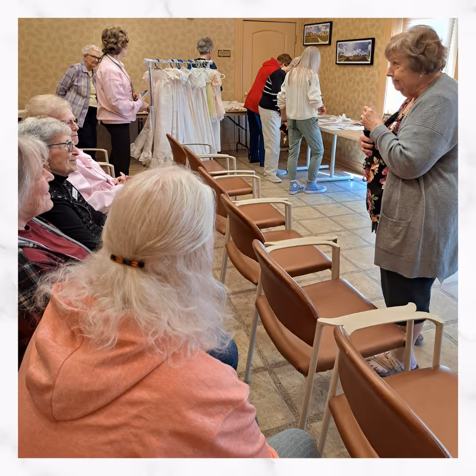 A group of elderly women gathered in a room with beige patterned wallpaper and tiled floor. Some women are seated in brown chairs arranged in rows, while others stand near a table with items on it and a clothing rack with white dresses. The atmosphere appears social and relaxed.
